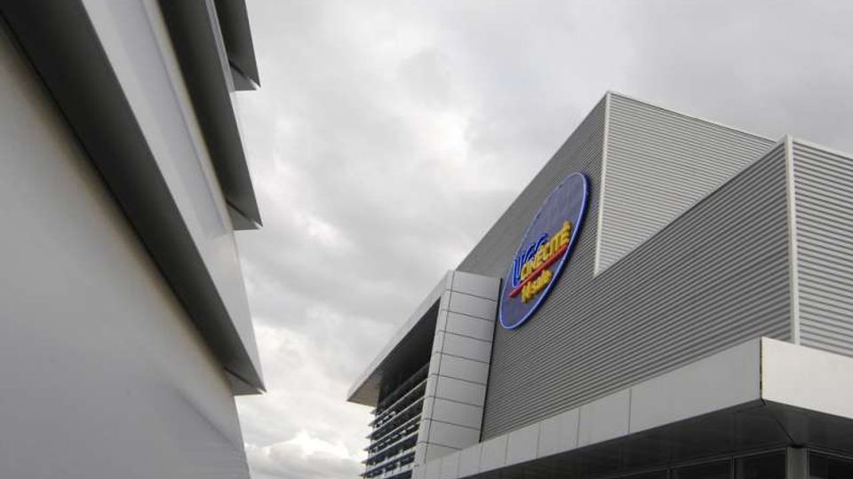 Edificio moderno con facciata in pannelli metallici grigi e ampie vetrate, con un grande logo ovale blu sulla parete. L’inquadratura dal basso mostra volumi angolari e una struttura adiacente, sotto un cielo nuvoloso.