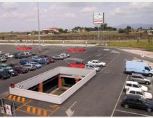 Ampio parcheggio asfaltato di un centro commerciale, con posti auto segnati, diverse vetture e aree con carrelli rossi; sullo sfondo si vedono un’insegna alta e una strada con campagna e colline lontane.