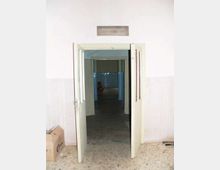 Ingresso interno con doppia porta a battente aperta che conduce a un corridoio stretto con pavimento in piastrelle scure e pareti in parte rivestite. In fondo si intravede un ambiente con piastrelle blu e luce più debole.