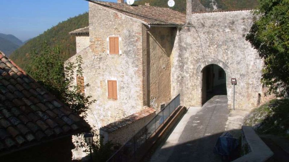Edificio in pietra e intonaco con tetto in coppi e finestre con persiane, affiancato a un grande arco che forma un passaggio coperto su una strada in salita. Sullo sfondo si vedono colline boscose e cielo sereno.