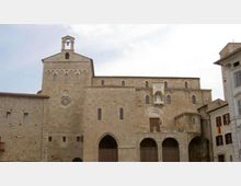 Facciata in pietra di una chiesa medievale con campanile a vela, rosone e finestrine ad arco; alla base si vedono portici con archi e grandi ingressi. L’edificio si affaccia su una piazza, con altri palazzi ai lati.