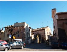 Piccola piazza in un borgo con pavimentazione in pietra e alcune auto parcheggiate, circondata da case basse in muratura con tetti in coppi. Al centro e sul lato destro si vedono scalinate che salgono tra gli edifici, con alberi potati e insegne stradali sotto un cielo sereno.