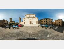 Ampia piazza lastricata in pietra con parcheggi e panchine, dominata al centro da una chiesa in stile neoclassico con facciata curva e portale principale. Intorno si vedono edifici storici a più piani e un campanile, sotto un cielo sereno.