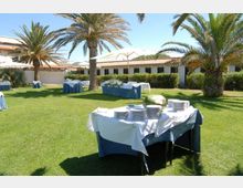 Giardino con prato verde e palme, allestito per un ricevimento con tavoli coperti da tovaglie bianche e blu, piatti impilati e ombrelloni. Sullo sfondo si vede un edificio basso con tetto in tegole e siepi lungo la facciata.