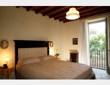 Camera da letto con grande letto matrimoniale e testata scura, soffitto con travi in legno e lampadario centrale. Ampia portafinestra con tenda chiara e balconcino con ringhiera, affacciata su alberi all’esterno.