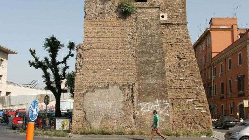 Torre medievale in mattoni con merlature e aperture ad arco, affiancata da resti di muratura e base scarpata. Si trova su un incrocio urbano con strisce pedonali, segnaletica stradale, alberi e palazzi moderni sullo sfondo.
