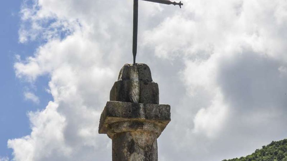 Croce metallica montata sulla sommità di una colonna in pietra all’aperto, ripresa contro un cielo azzurro con nuvole. Sullo sfondo si vede un pendio montano coperto di vegetazione verde e un angolo di edificio in pietra.