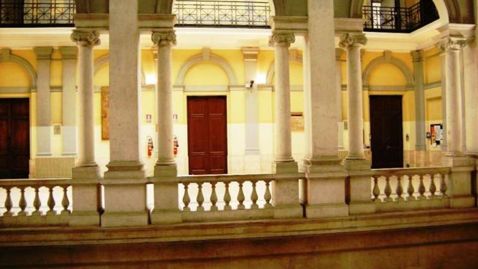 Cortile interno di un edificio storico con porticato su due livelli, grandi archi e colonne in pietra. Al piano superiore si vedono balaustre e ringhiere in ferro, con pareti color crema e porte scure sullo sfondo.