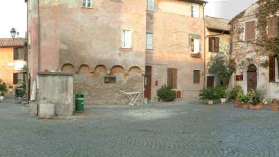 Piazza lastricata in pietra con edifici storici in muratura dai toni rosati e ocra, finestre con persiane e piccoli archi alla base. Ai bordi si vedono vasi con piante e un edificio parzialmente coperto da vegetazione, con ingressi e dettagli architettonici tradizionali.