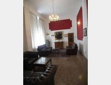 Ampio salone con soffitto alto, lampadario centrale, grandi finestre con tende chiare e pareti bianche con riquadri decorativi rossi. L’ambiente è arredato con divani e poltrone in pelle scura, tavolini, tappeto e una credenza con specchio e quadri alle pareti.