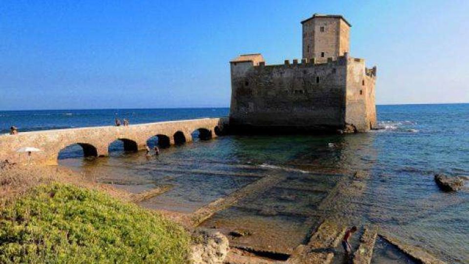 Castello costiero in pietra su un isolotto, collegato alla riva da un ponte basso ad arcate sopra il mare poco profondo. In primo piano si vedono una piccola spiaggia rocciosa con resti di strutture rettangolari e vegetazione, sullo sfondo il mare aperto e il cielo sereno.