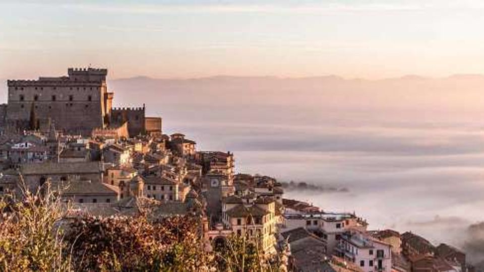 Borgo collinare con case in pietra addossate tra loro e un grande castello fortificato in cima. In lontananza si vedono colline e una valle coperta da nebbia sotto un cielo chiaro.
