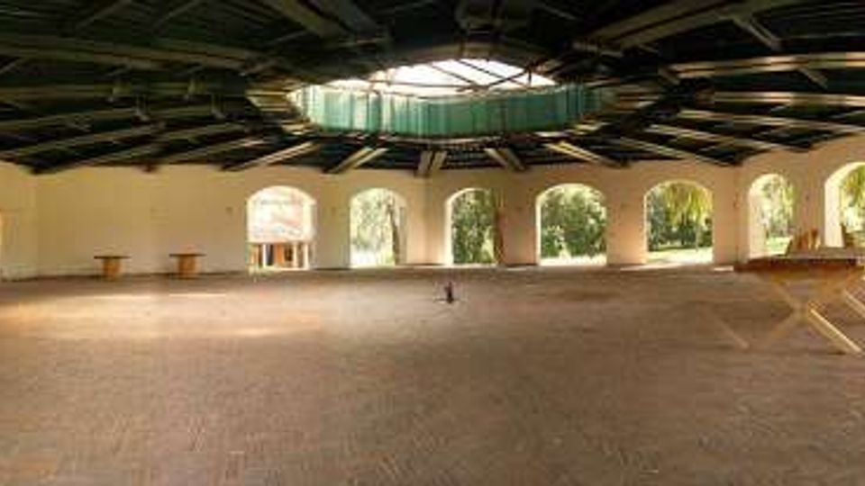 Ampio padiglione aperto con pavimento in mattoni e grandi arcate lungo il perimetro, affacciate sul verde esterno. Tetto alto con travi a vista e lucernario centrale che illumina lo spazio interno.