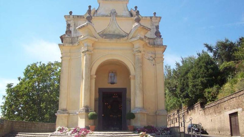 Facciata di una piccola chiesa in stile barocco, color crema, con portico ad arco, colonne e croce in sommità. Davanti, una scalinata in pietra con aiuole fiorite conduce all’ingresso, circondato da alberi e un muro laterale.