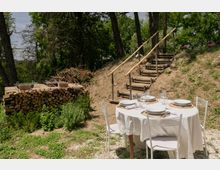 Tavolo rotondo all’aperto apparecchiato con tovaglia bianca, piatti e calici su un’area di ghiaia, circondato da verde. Sullo sfondo si vedono una scalinata in tronchi con corrimano in legno su un terrapieno, alberi e una catasta di legna con griglia.