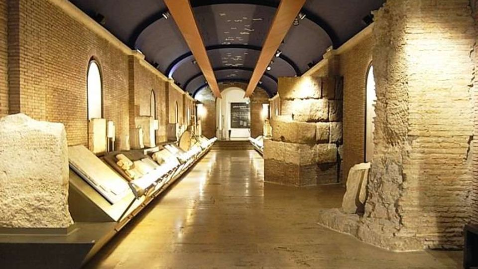Interno di un museo archeologico con lunga galleria in pietra e mattoni, vetrine espositive illuminate lungo le pareti e grandi blocchi di rovine ai lati. Soffitto ad arco scuro con travi e luci incassate, pavimento in lastre di pietra con targhe e segnalazioni.