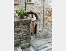 Vicolo in pietra in un borgo storico, con una scalinata acciottolata che scende sotto un arco in muratura collegato a una parete in pietra grezza. Si vede una piccola porta ad arco con cornice decorata e un muretto in pietra con vaso di piante.