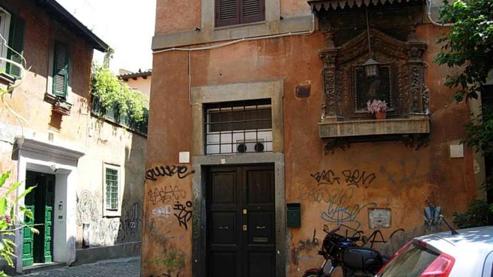 Facciata di un edificio color ocra su una stretta strada acciottolata, con portone nero, finestra con inferriata e graffiti sui muri. A destra sporge un piccolo balconcino decorato con edicola e lampada, con una moto parcheggiata e parte di un’auto in primo piano.