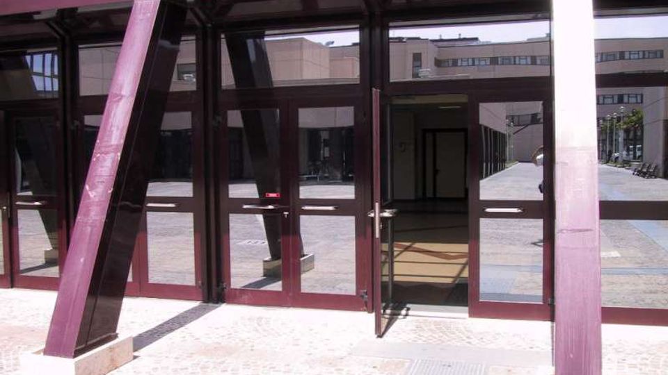 Ingresso di un edificio moderno con grandi vetrate e telai metallici inclinati color bordeaux, con porte in vetro che si aprono su un atrio interno. Davanti si vede una pavimentazione in pietra e, sullo sfondo, un cortile con altri edifici.