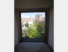 Vista attraverso una piccola finestra quadrata con cornice in cemento, da un interno spoglio, su un’area verde con alberi e palme. Sullo sfondo si vedono parcheggio e palazzine residenziali a più piani sotto un cielo parzialmente nuvoloso.