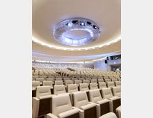 Ampia sala auditorium moderna con molte file di poltrone beige disposte a gradoni. Soffitto curvo con grande elemento circolare illuminato al centro e luci incassate lungo le pareti.