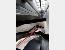 Interno di una scala moderna in cemento a più rampe con parapetti neri curvi, corrimano rosso e gradini in griglia metallica. In alto un lucernario longitudinale illumina l’ampio vano verticale.