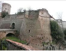 Ruderi di una fortificazione in muratura con grande bastione in pietra e torre cilindrica sullo sfondo. In primo piano si vede un arco di ponte e un tetto in coppi, con vegetazione e pareti rocciose attorno alle mura.