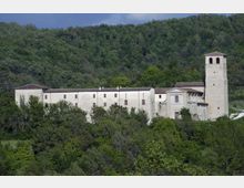 Grande complesso monastico in pietra chiara con edifici allungati e una torre campanaria quadrata, situato ai margini di una collina boscosa. La struttura è circondata da vegetazione fitta e si staglia sullo sfondo di un pendio verde.
