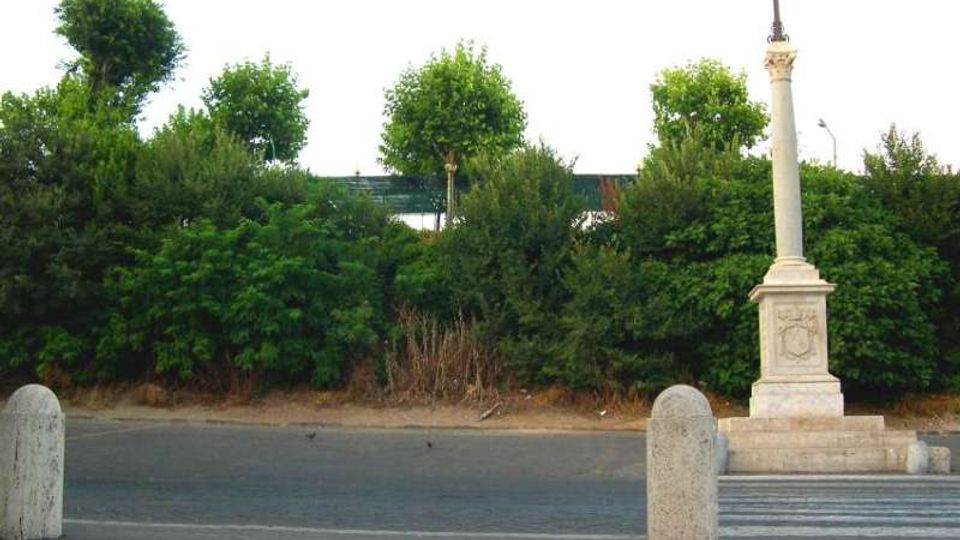 Piazzale asfaltato con cordolo in pietra e alcuni paracarri, davanti a una fitta fascia di vegetazione e alberi. A destra si trova un monumento in pietra con basamento e colonna sormontata da una croce.