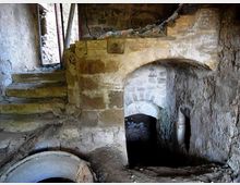 Interno di un ambiente sotterraneo in pietra e mattoni, con arco ribassato che conduce a un passaggio buio e una breve scala laterale in muratura. Si vedono pareti grezze, detriti sul pavimento e una grande apertura circolare in primo piano.