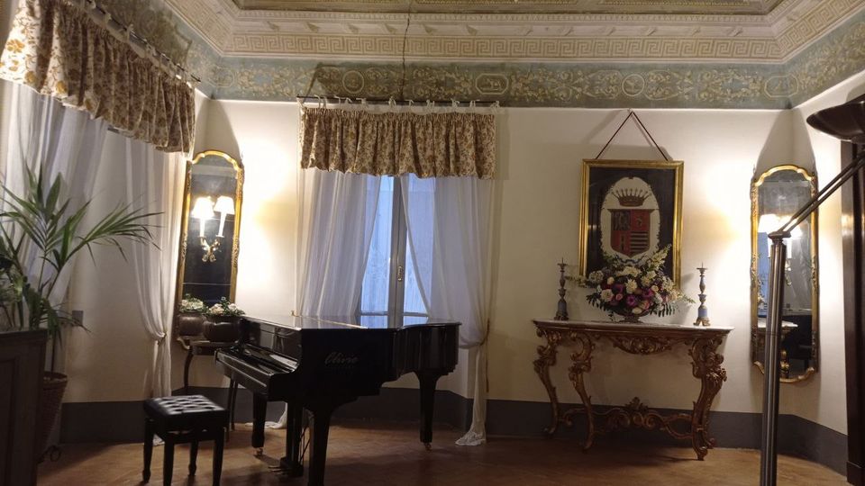 Salone interno in stile classico con soffitto affrescato e cornici decorative, pavimento in cotto e grandi finestre con tende bianche e mantovane floreali. Al centro un pianoforte a coda nero, accanto a una consolle intagliata con composizione floreale, specchi con appliques e un quadro con stemma sulla parete.