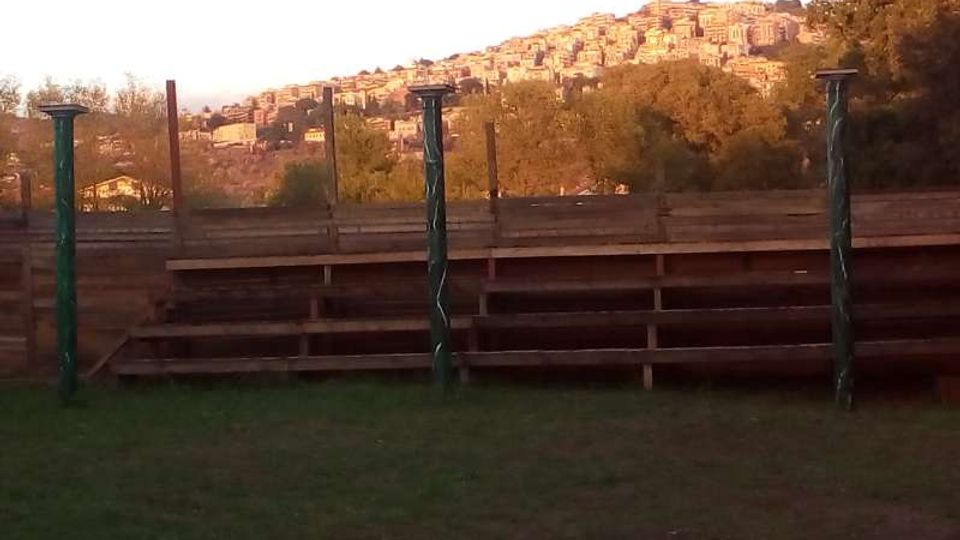 Piccole gradinate in legno all’aperto su un prato, con pali verticali lungo il perimetro. Sullo sfondo si vede una collina con molte case addossate tra alberi e vegetazione.
