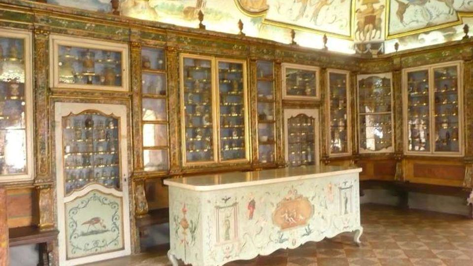 Interno di una sala storica con pareti rivestite da grandi vetrine e armadi dorati pieni di piccoli oggetti esposti. Al centro c’è un banco decorato, mentre il soffitto è affrescato con motivi ornamentali e figure.