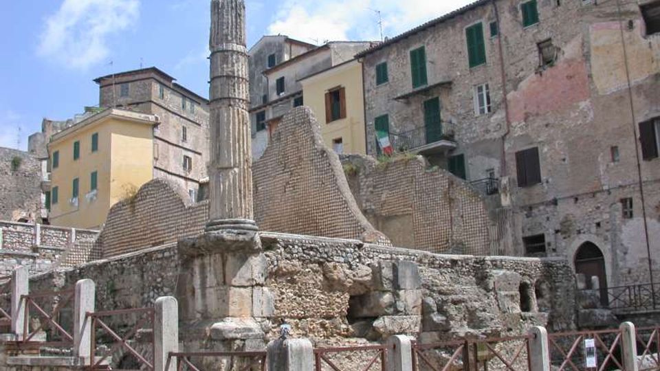 Rovine archeologiche in pietra con una colonna antica isolata e muri parzialmente crollati, circondate da una recinzione metallica. Sullo sfondo si vedono edifici storici in muratura con persiane verdi affacciati sulla piazza sotto un cielo parzialmente nuvoloso.