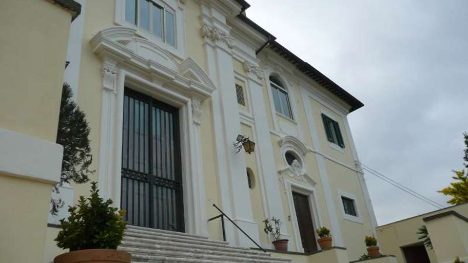 Facciata di un edificio storico in stile barocco, intonacata in giallo chiaro con stucchi bianchi, portale alto con inferriata e finestre ad arco. Davanti, una scalinata in pietra con corrimano e vasi di piante conduce all’ingresso sotto un cielo nuvoloso.