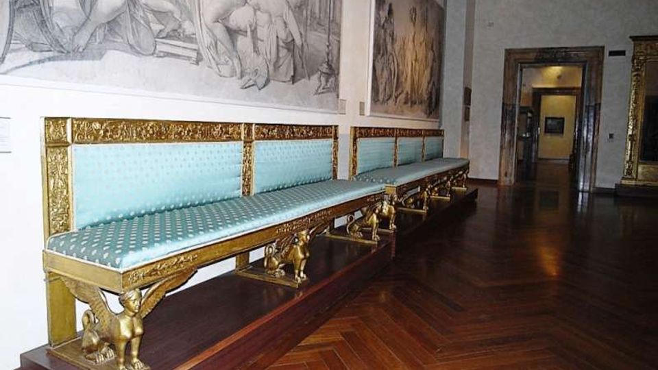 Sala museale con lungo divano imbottito azzurro e decorazioni dorate, sostenuto da piedini scolpiti, su pavimento in parquet a spina di pesce. Alle pareti sono visibili grandi disegni murali e, in fondo, un passaggio ad arco verso un’altra stanza.
