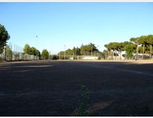 Ampio campo sportivo all’aperto con fondo scuro in terra battuta, circondato da recinzioni metalliche e pali per l’illuminazione. Sullo sfondo si vedono alberi e una piccola struttura vicino ai campi, sotto un cielo sereno.