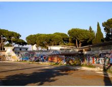 Ampio cortile asfaltato circondato da edifici bassi con tettoie e lunghe pareti ricoperte di graffiti colorati. Sullo sfondo si vedono grandi pini e cipressi sotto un cielo sereno.