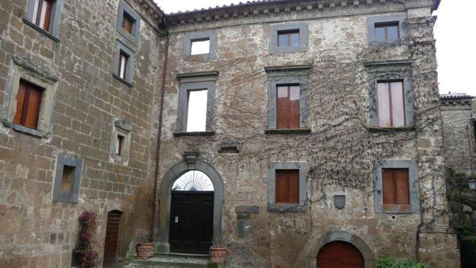 Cortile interno di un edificio storico in pietra con facciata a più piani, finestre con scuri in legno e archi in muratura. Si vedono un grande portone ad arco con gradini in pietra, un accesso ad arco più piccolo e pareti parzialmente coperte da rampicanti.