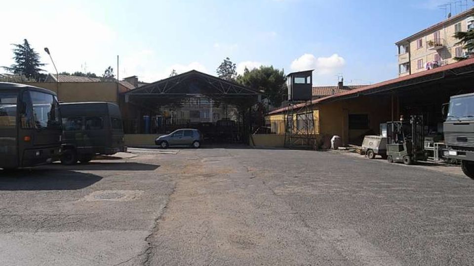 Ampio piazzale asfaltato di un’area industriale con capannoni e tettoie laterali, alcuni camion e veicoli parcheggiati. Sullo sfondo si vede un grande ingresso coperto con struttura metallica e un edificio residenziale a più piani sulla destra.