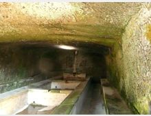 Interno di una vasca coperta in pietra, con soffitto basso e pareti umide ricoperte di muschio. Al centro corre un lungo canale in muratura con comparti rettangolari, illuminato debolmente da una piccola apertura superiore.