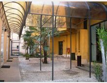 Cortile interno di un edificio con pavimento in ghiaia e piccole palme al centro, circondato da muri gialli con porte e finestre. Una tettoia arcuata in metallo e pannelli traslucidi copre il passaggio laterale e parte dello spazio, con pali verticali e un cancello sul fondo.