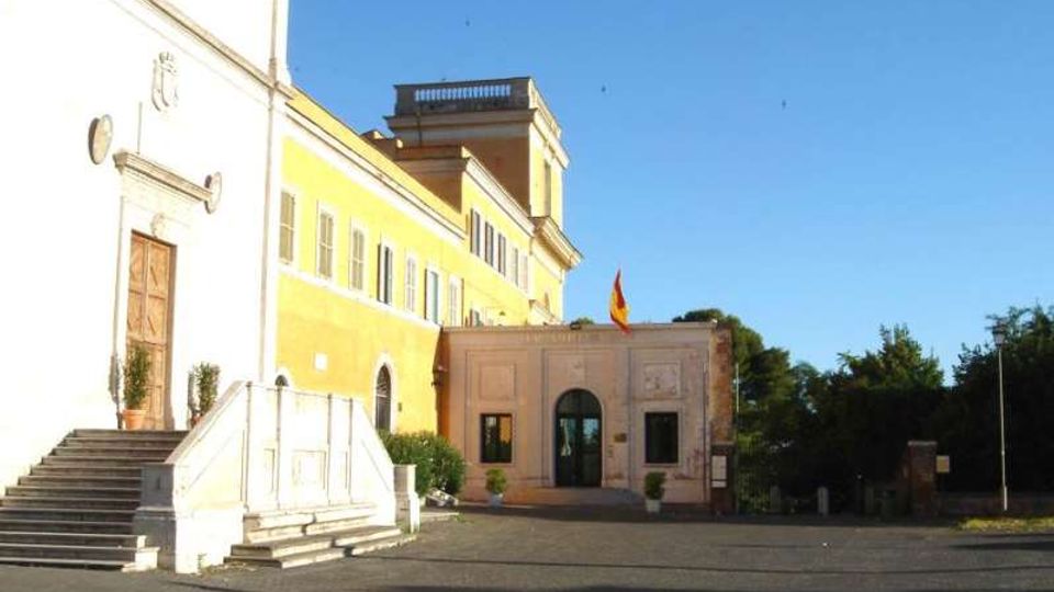 Edificio istituzionale con facciata gialla e bianca, scalinata d’ingresso in pietra e portale laterale, affacciato su un ampio piazzale asfaltato. Al centro si vede un’entrata ad arco con bandiera su un corpo più basso, con alberi e vegetazione sullo sfondo sotto un cielo sereno.