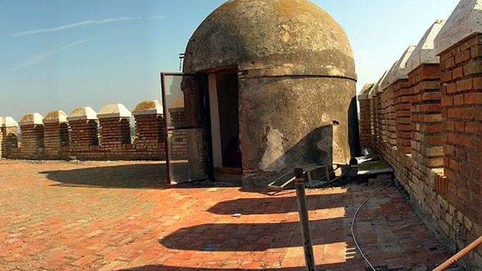 Terrazza in mattoni su una fortificazione con parapetti merlati e un piccolo edificio a pianta circolare con cupola in pietra e porta aperta. Lo spazio è all’aperto, affiancato da muri in mattoni e con vista sul cielo limpido.