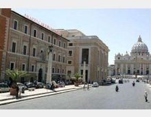 Ampia piazza urbana con strada asfaltata e palazzi storici a più piani con facciate in pietra e finestre regolari; sullo sfondo si vede la Basilica di San Pietro con la grande cupola. Ai lati sono presenti lampioni, alcune palme in vaso e auto parcheggiate.