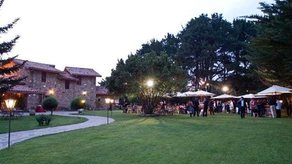 Ampio prato con vialetto curvo e lampioni che conduce a un casale in pietra con tetto in tegole. Sullo sfondo si vedono grandi alberi e un’area esterna con ombrelloni bianchi e illuminazione serale.