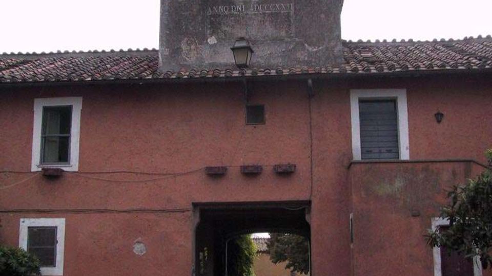 Facciata di un edificio storico intonacato color rosso con tetto in coppi e passaggio ad arco centrale che conduce a un cortile interno. Sopra l’ingresso si alza una torre con grande orologio e due campanili gemelli con campane, sormontati da croci.