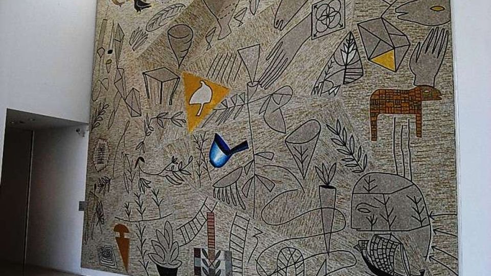 Ampio atrio interno moderno con pavimento in pietra grigia, pareti bianche e finestre alte a nastro vicino al soffitto. Una grande parete è occupata da un murale astratto su fondo beige con figure geometriche e motivi di foglie e animali stilizzati.