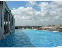 Piscina a sfioro su una terrazza panoramica, affiancata da una struttura con vetrate e tende bianche. Sullo sfondo si vedono tetti e edifici della città sotto un cielo parzialmente nuvoloso.