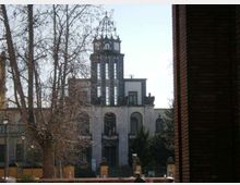 Edificio storico con facciata chiara e una torre centrale alta con finestre verticali e orologio, sormontata da una struttura metallica. In primo piano si vedono alberi spogli e una recinzione, con un muro in mattoni sul lato destro dell’inquadratura.